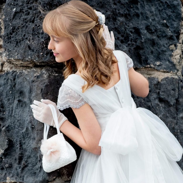 Niña con vestido blanco de comunión, peinado con moño, sosteniendo un bolso y mirando hacia un lado.