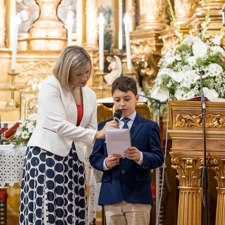 Niño leyendo un discurso en un evento religioso, acompañado por una mujer. Decoración floral al fondo.